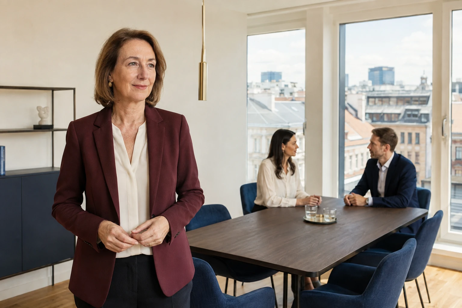 Frau in hellem Besprechungsraum, zwei Personen am Tisch, städtische Dächer draußen.